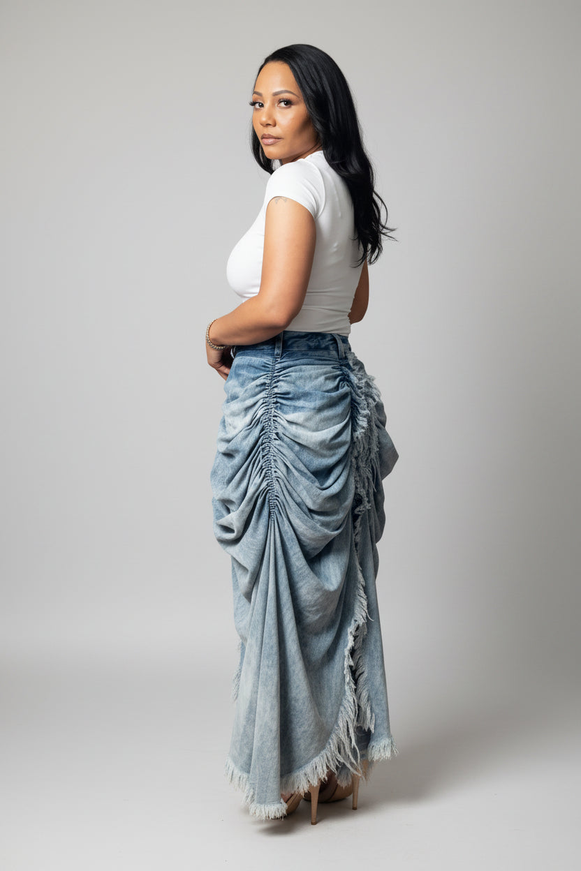 A woman shown from the back wearing a white short-sleeve top and a long, gathered light blue denim skirt with fringe detailing, set against a plain white background.