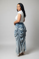 A woman shown from the back wearing a white short-sleeve top and a long, gathered light blue denim skirt with fringe detailing, set against a plain white background.