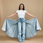 A woman stands in a doorway wearing a fitted white short-sleeve T-shirt and high-waisted light blue wide-leg jeans with a matching light blue tie-dye shirt draped over her lap. Behind her is a white wall with two framed line drawings and a closet shelf filled with shoes.