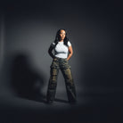A woman stands in a doorway of a home interior, wearing a white short-sleeve top and distressed wide-leg cargo pants in a faded olive-brown color. To her left is a white wall with two framed black-and-white sketches, and behind her is an open closet filled with shoes.