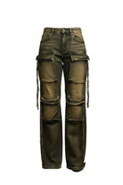 A woman with a tattoo on her left arm stands in a doorway of a home, wearing a white short-sleeve t-shirt and distressed olive green cargo pants with fringe details. Behind her is an open closet filled with shoes, and to her left are two framed black and white sketches on a white wall.